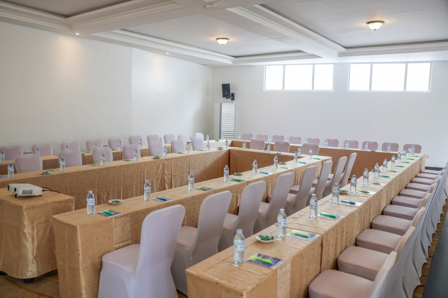 Long tables in classroom rows with covered chairs, notepads and water set out.