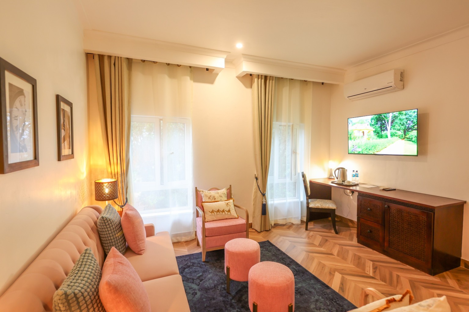 A Deluxe Room with sofa and ottomans, writing desk, wall-mounted television and a window onto the garden.