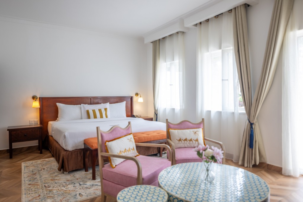 A Deluxe Room made up with twin beds, a pair of armchairs and a small round table beside the curtained window.