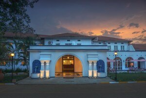 The hotel’s white 1950s facade at dusk, its central arch lit gold beneath a violet sky.