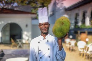 A chef in whites holding up a whole jackfruit on the terrace.