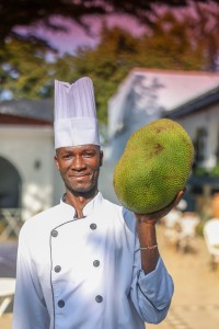 A chef in whites holding up a whole jackfruit on the terrace.