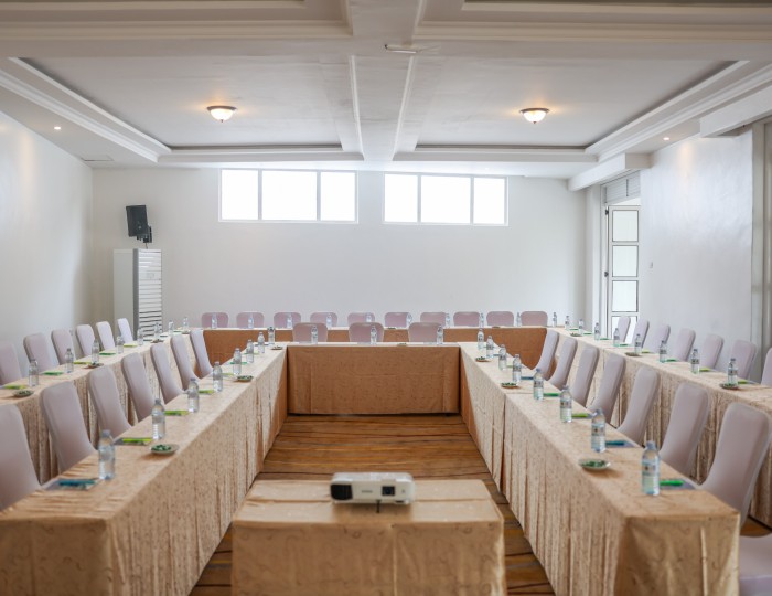 A conference hall in a U-shaped layout under daylight windows, set with notepads, water and a projector.