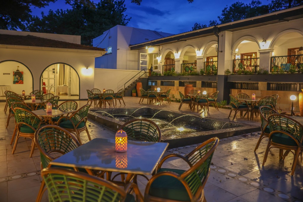 Courtyard tables and cane chairs set around a lit water feature at dusk, the arcades glowing behind them.