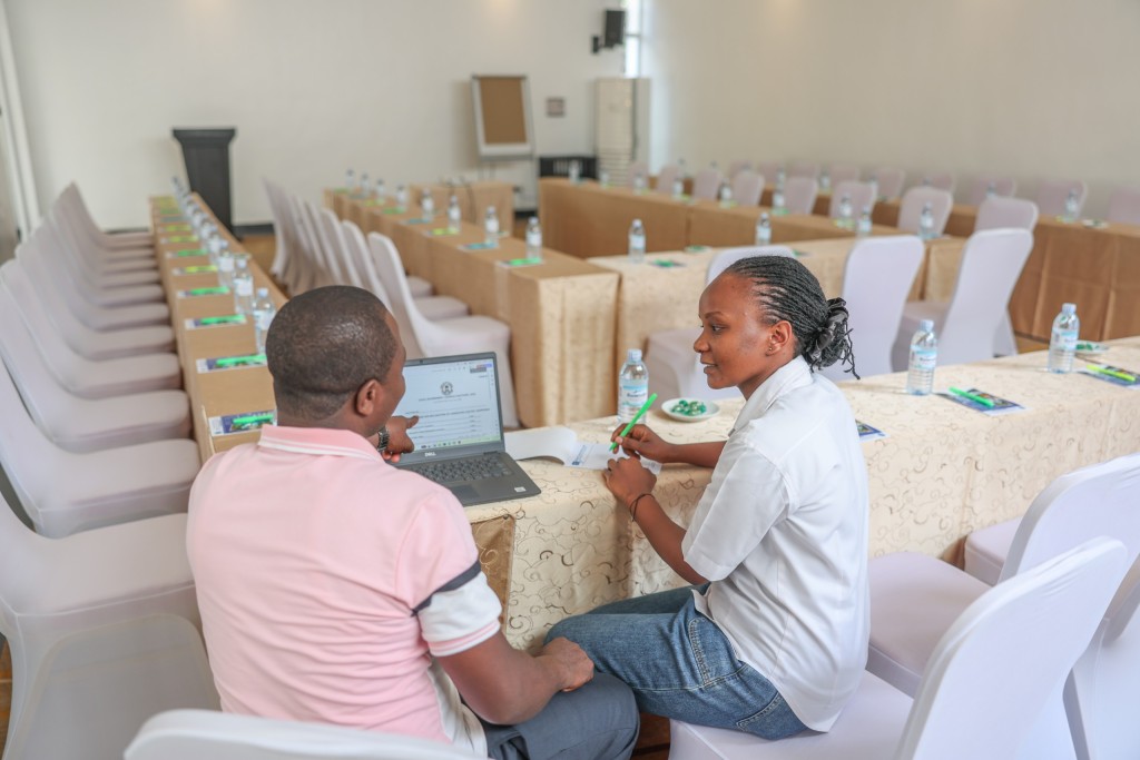 A delegate working at a laptop at a skirted classroom table, a colleague beside him and further rows behind.