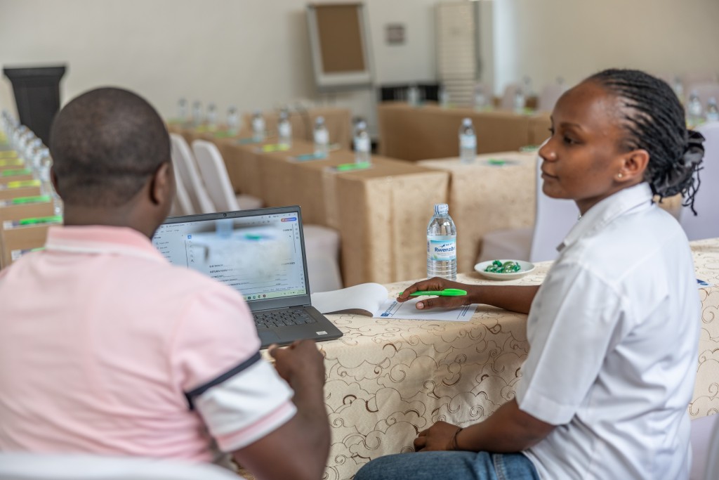 A participant working at a laptop at a skirted table with a colleague beside him and further rows behind.