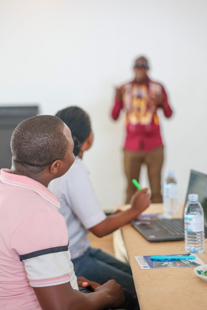 Two delegates seated at a skirted table with a laptop and notes, a facilitator standing at the front beyond them.