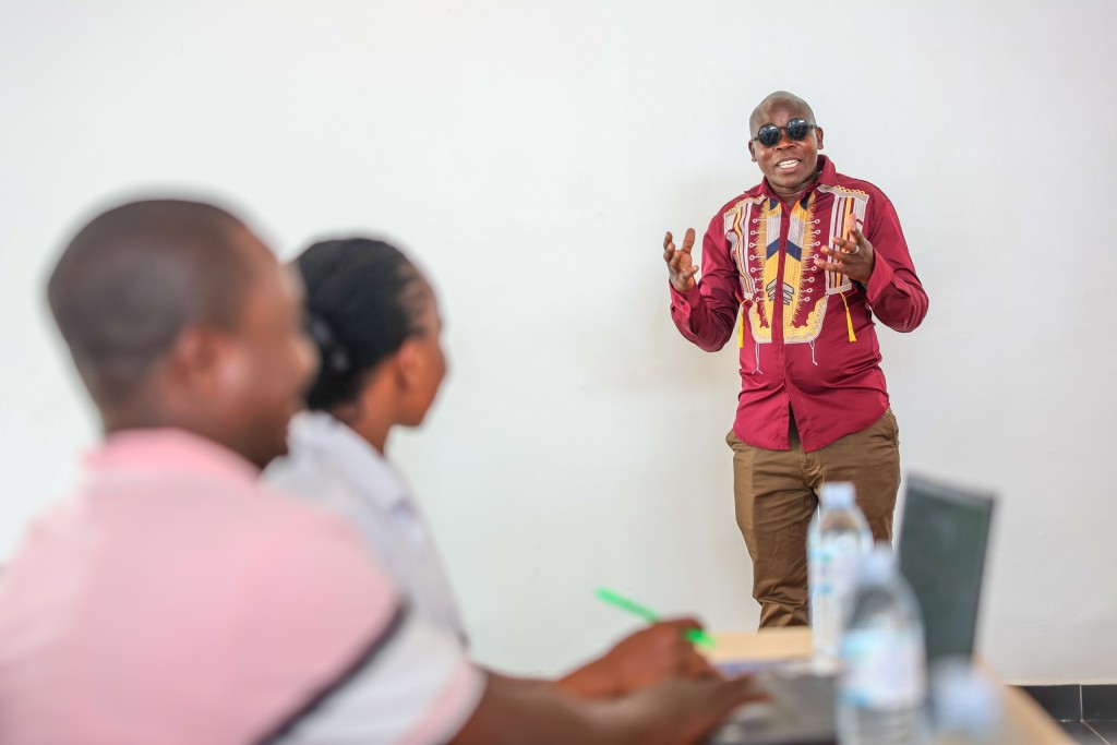 A facilitator presenting at the front of the meeting room, two delegates in the foreground taking notes.