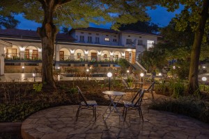 The lit arcaded frontage of the hotel at dusk, seen across the lawns and garden paths.