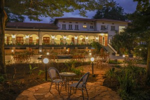 The lit arcaded frontage of the hotel at dusk, seen across the lawns and garden paths.