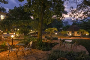 The lit arcaded frontage of the hotel at dusk, seen across the lawns and garden paths.