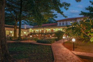 The lit arcaded frontage of the hotel at dusk, seen across the lawns and garden paths.
