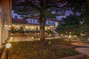 The lit arcaded frontage of the hotel at dusk, seen across the lawns and garden paths.