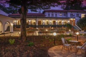 The lit arcaded frontage of the hotel at dusk, seen across the lawns and garden paths.