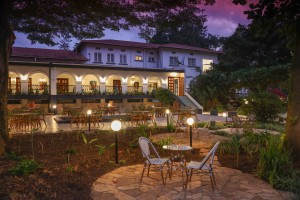 The lit arcaded frontage of the hotel at dusk, seen across the lawns and garden paths.