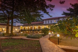 The lit arcaded frontage of the hotel at dusk, seen across the lawns and garden paths.