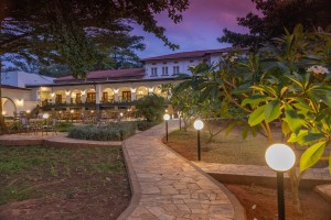 The lit arcaded frontage of the hotel at dusk, seen across the lawns and garden paths.