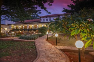 The lit arcaded frontage of the hotel at dusk, seen across the lawns and garden paths.