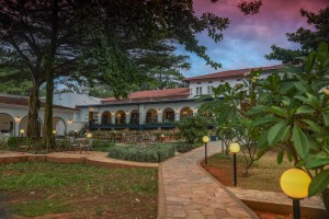 The lit arcaded frontage of the hotel at dusk, seen across the lawns and garden paths.