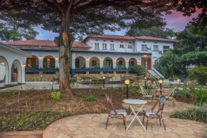 The lit arcaded frontage of the hotel at dusk, seen across the lawns and garden paths.
