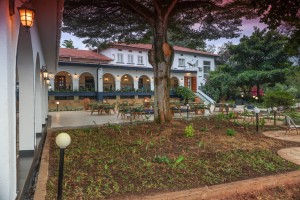 The lit arcaded frontage of the hotel at dusk, seen across the lawns and garden paths.