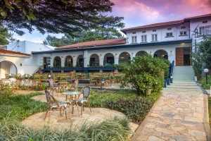 The lit arcaded frontage of the hotel at dusk, seen across the lawns and garden paths.