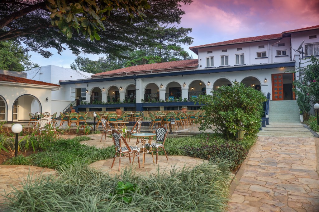 Garden chairs and tables spread across the lawn below the hotel’s lit arcades at dusk, with steps up to the terrace.