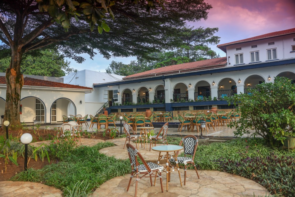 Garden tables under a mature tree, with the hotel’s lit arcaded frontage behind them at dusk.