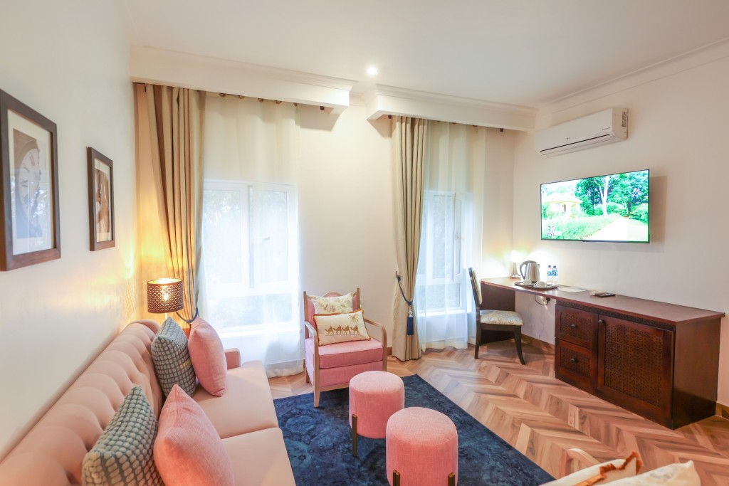 A guest room with a sofa and ottomans, a timber writing desk under a wall-mounted television and a kettle tray.