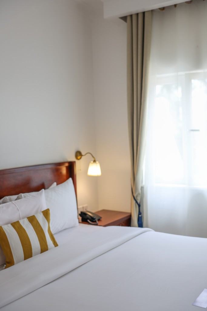 A made-up bed beside a curtained window, daylight falling across white linen and a reading lamp.