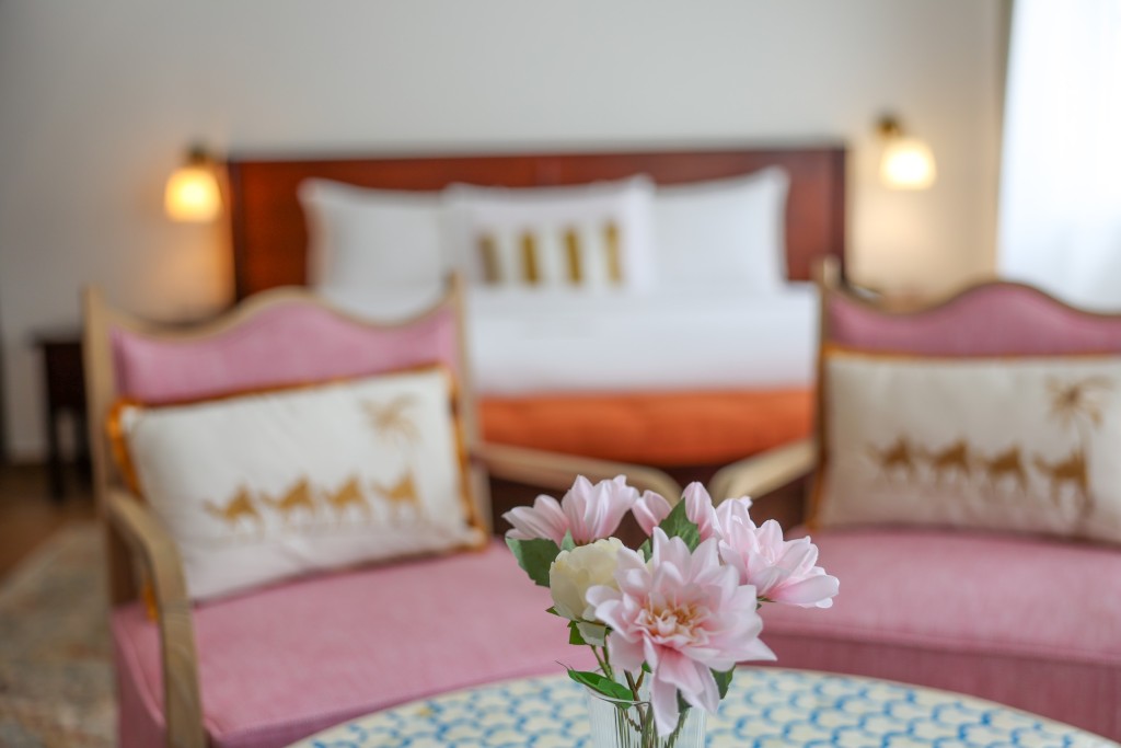 Twin beds made up side by side, flowers on the patterned table in front of them and lamps lit either side.