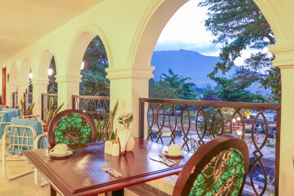 A laid table on the restaurant veranda, looking out through a tall arch to a wooded ridge beyond the garden.