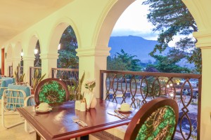 Masaaba Restaurant: tables and cane chairs along the arched veranda, hills and greenery through the openings.