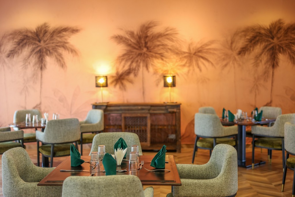 A warm terracotta dining room painted with tall palms, its tables laid with green napkins under low wall lamps.