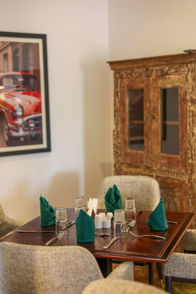 A two-place table laid with green napkins, a carved timber cabinet and a framed print on the wall behind.