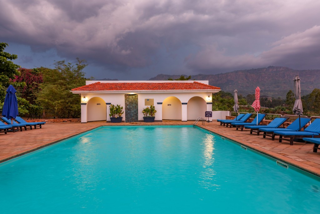 The swimming pool at dusk under a heavy sky, the pool pavilion lit at the far end and hills behind it.