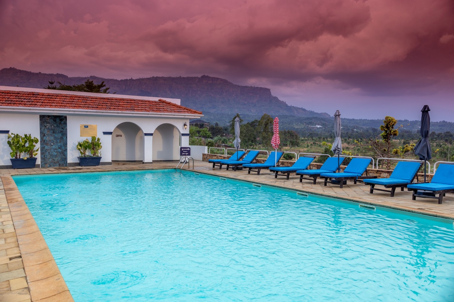 The hotel swimming pool at sunset, a row of blue sun loungers along the far side and the Mount Elgon escarpment ridge behind the arched pool pavilion.