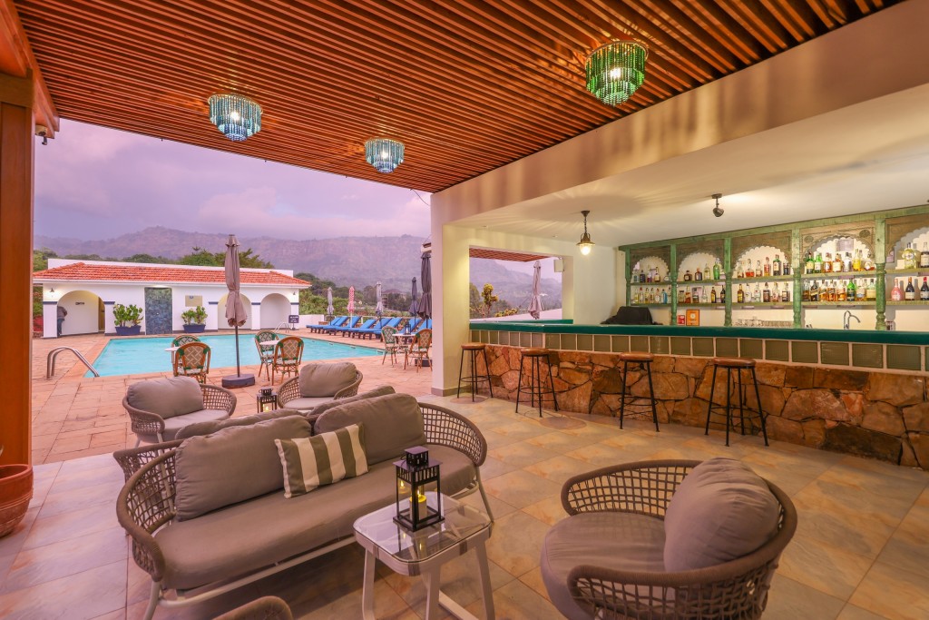 The open-sided poolside bar, stools at its stone counter and cane lounge seating, looking out across cafe tables to the pool and the hills beyond.