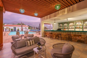 The open-sided poolside bar, stools at its stone counter and cane lounge seating, looking out across cafe tables to the pool and the hills beyond.