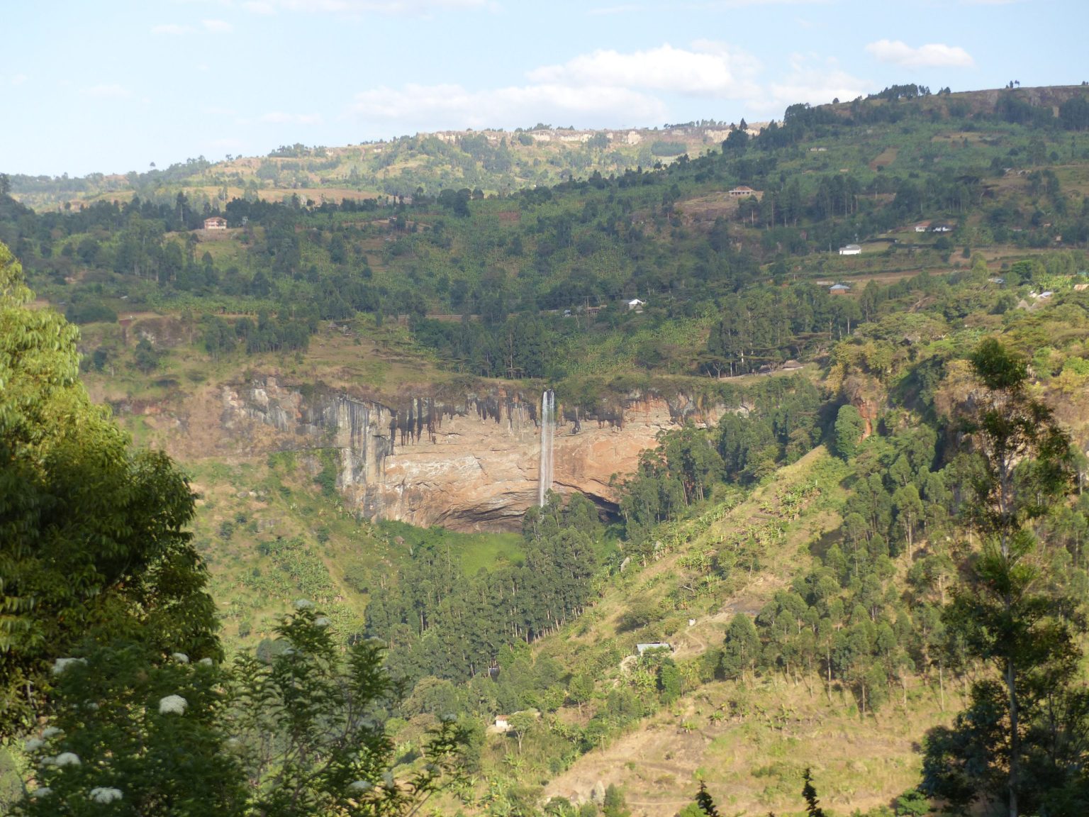 Sipi Falls seen from across the valley, dropping over a cliff among farmland and trees.