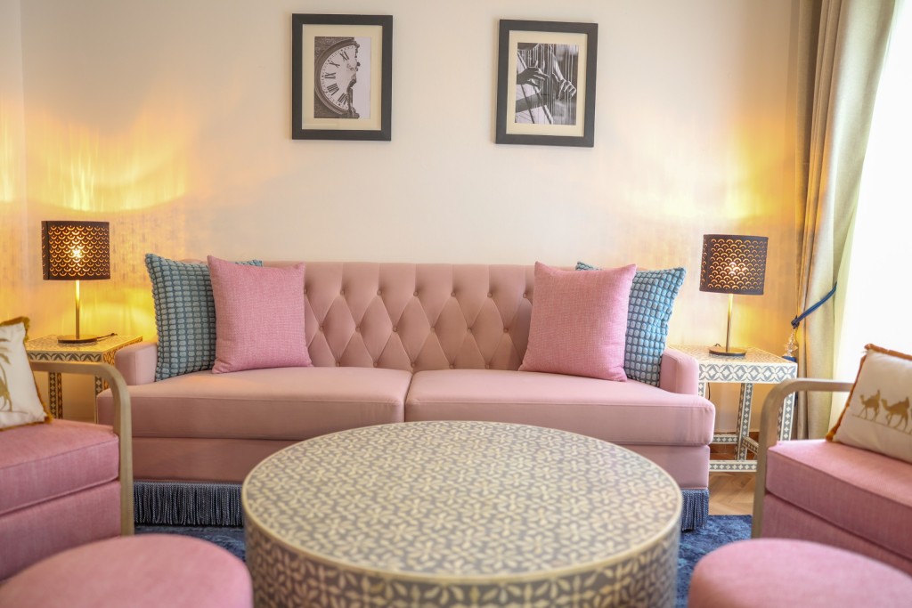 A suite sitting area with a pink buttoned sofa, armchairs and a patterned round table beneath framed prints.